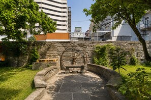 Ein Stebrunnen mit einer hölzernen Bank und einem Rad, umgeben von Gras und Bäumen, mit modernen Gebäuden im Hintergrund.