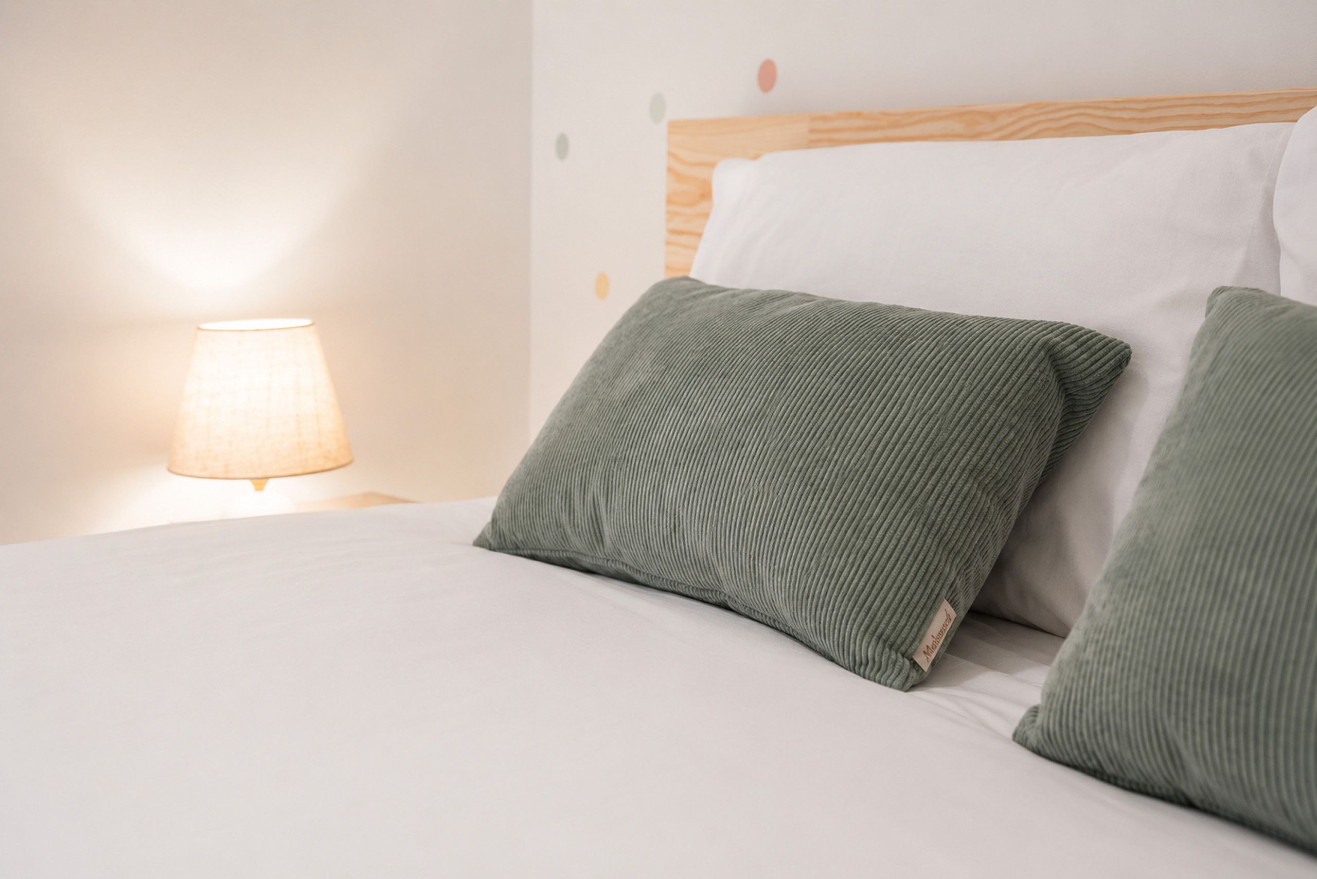 Ein Bett mit weißer Bettwäsche und zwei grünen Kissen, eine Lampe auf dem Nachttisch und ein hölzernes Kopfteil.