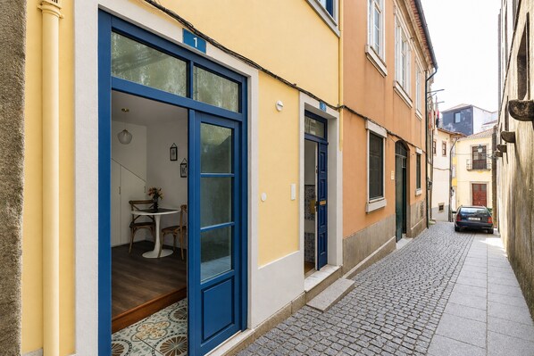 Una calle estrecha flanqueada por edificios, con una puerta azul y una pequeña mesa con sillas frente a un inmueble.