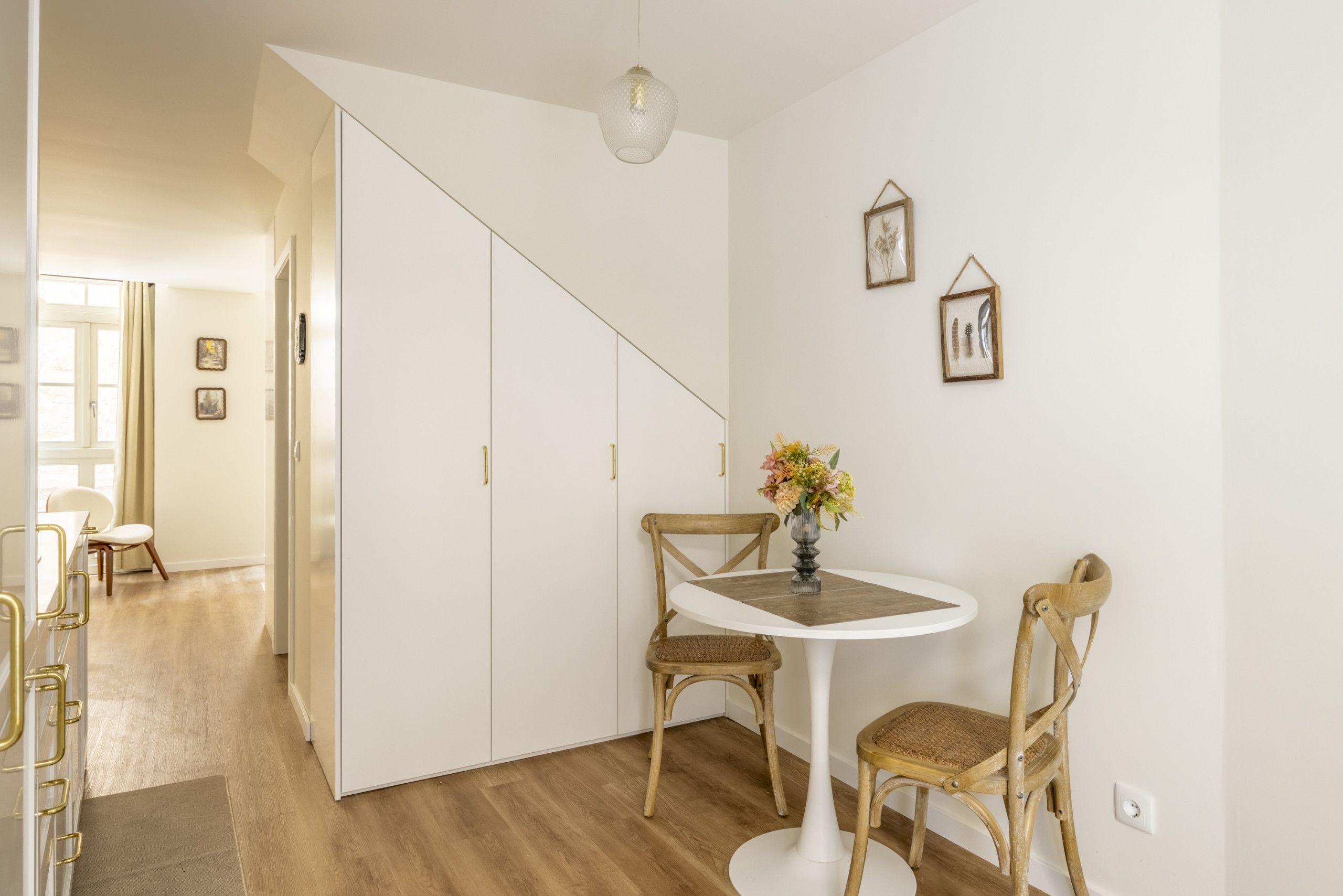 Une pièce avec une table blanche, deux chaises, un vase de fleurs et un escalier menant à une autre pièce.