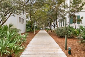 A wooden pathway flanked by greenery and white buildings with green shutters. - EZ Walk to Camp WaterColor! LSV/Golf Cart & 4 Bikes Included! (Santa Rosa Beach)