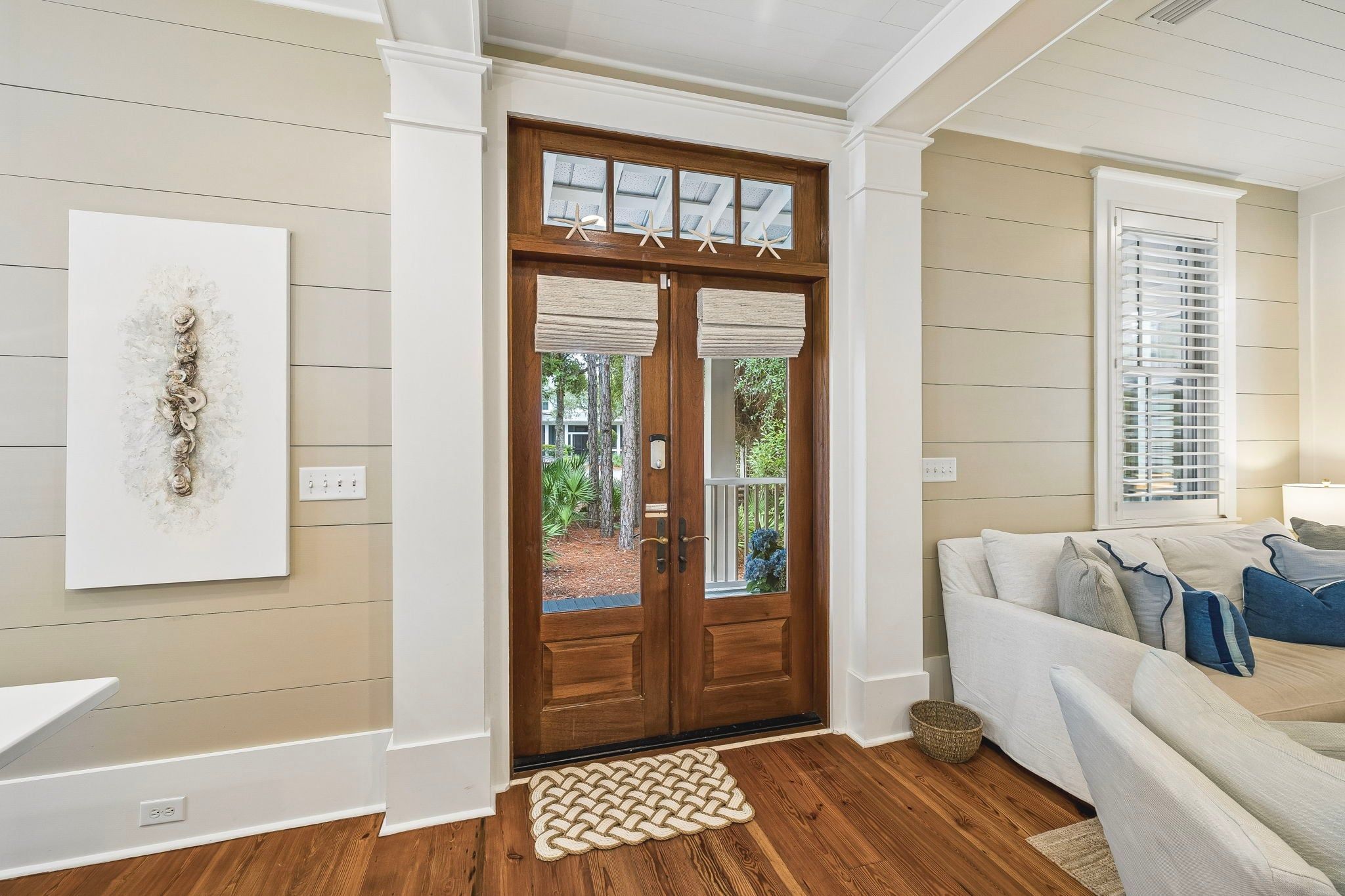 A room with a wooden door, a white wall with horizontal lines, a white sofa with cushions, and a woven rug.