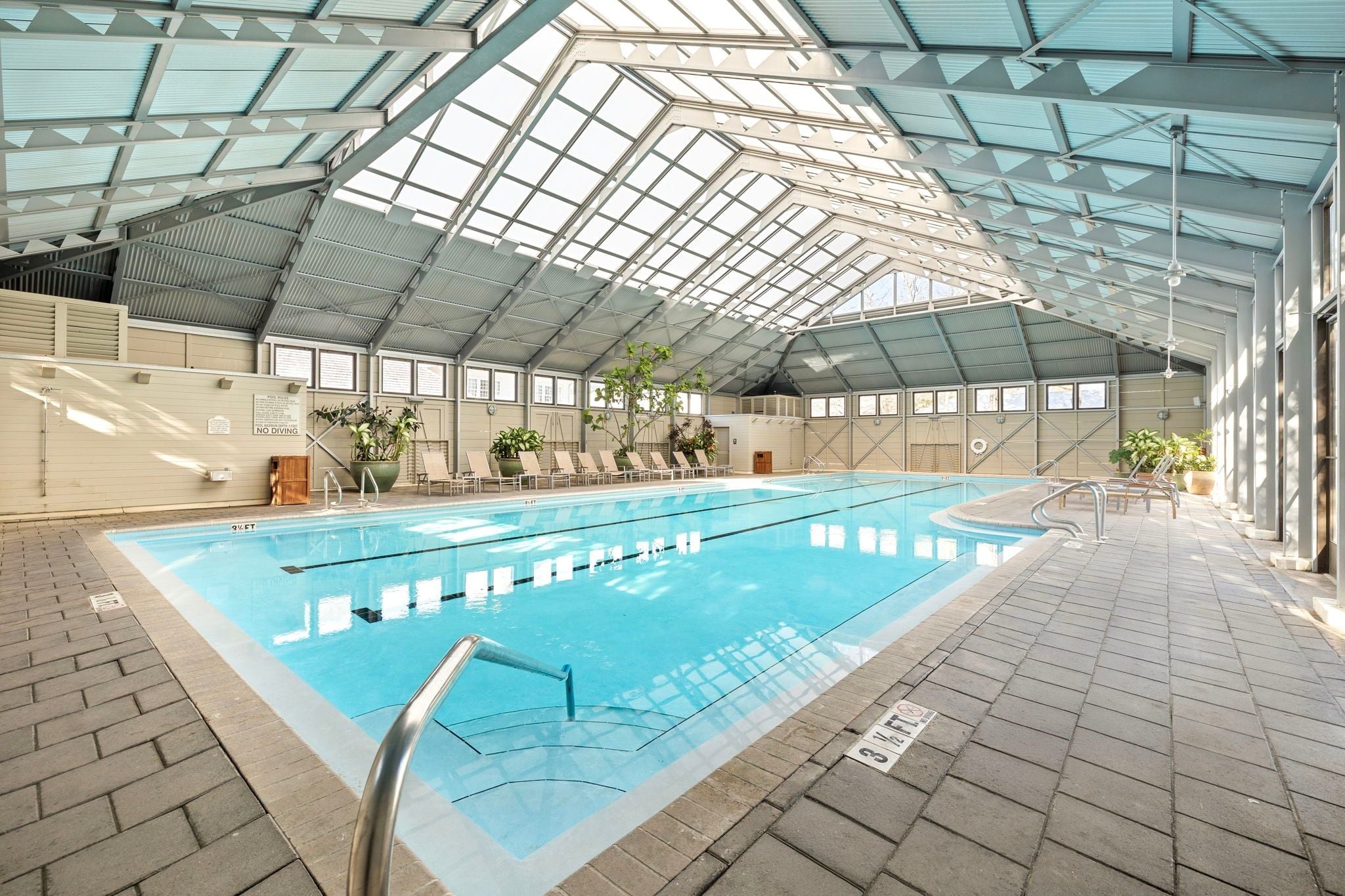 Une piscine intérieure dotée d’un haut plafond, de panneaux de verre transparent et de chaises longues disposées le long des parois.