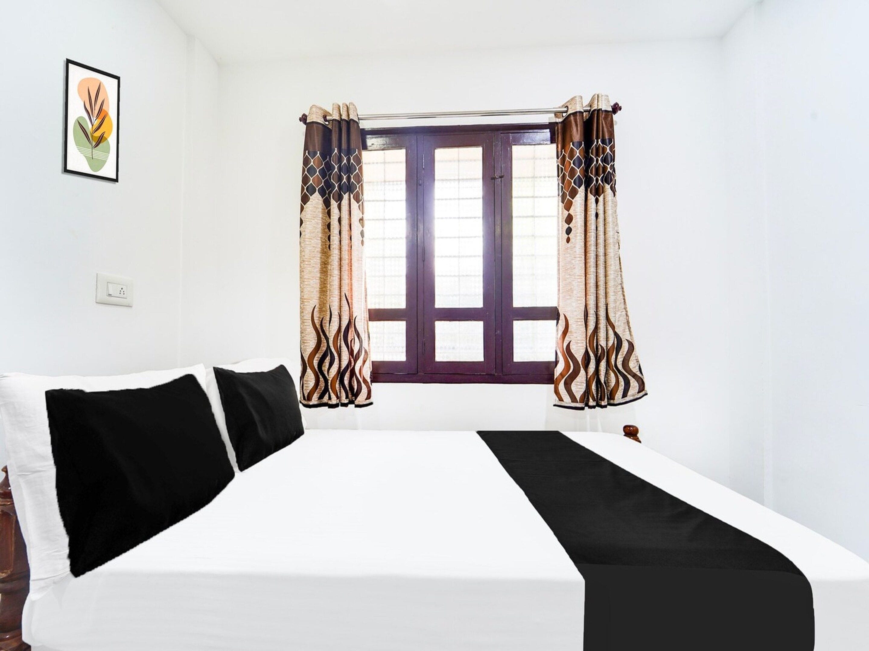 Una habitación con cama, una ventana con cortinas, un interruptor de luz en la pared y un cuadro enmarcado.