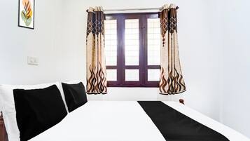 Una habitación con cama, una ventana con cortinas, un interruptor de luz en la pared y un cuadro enmarcado.