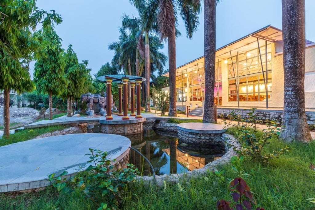 A modern building with a glass facade, surrounded by a landscaped garden with palm trees and a small pond.