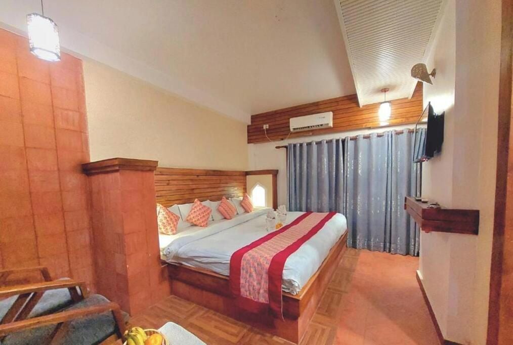 A hotel room with a wooden headboard, a bed with a red and white bedspread, a flat-screen TV mounted on the wall, and a window with curtains.