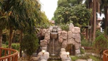 A stone bridge over a pond with a waterfall, surrounded by trees and a fence.