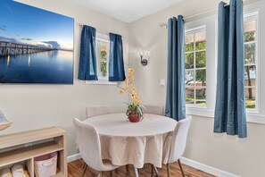 A dining area with a round table, chairs, a vase with flowers, a painting on the wall, and curtains on the windows. - Family‑Friendly 2BR Beach Home • Walk to Beach & Shops 🌴🏖️ (Cape Canaveral)
