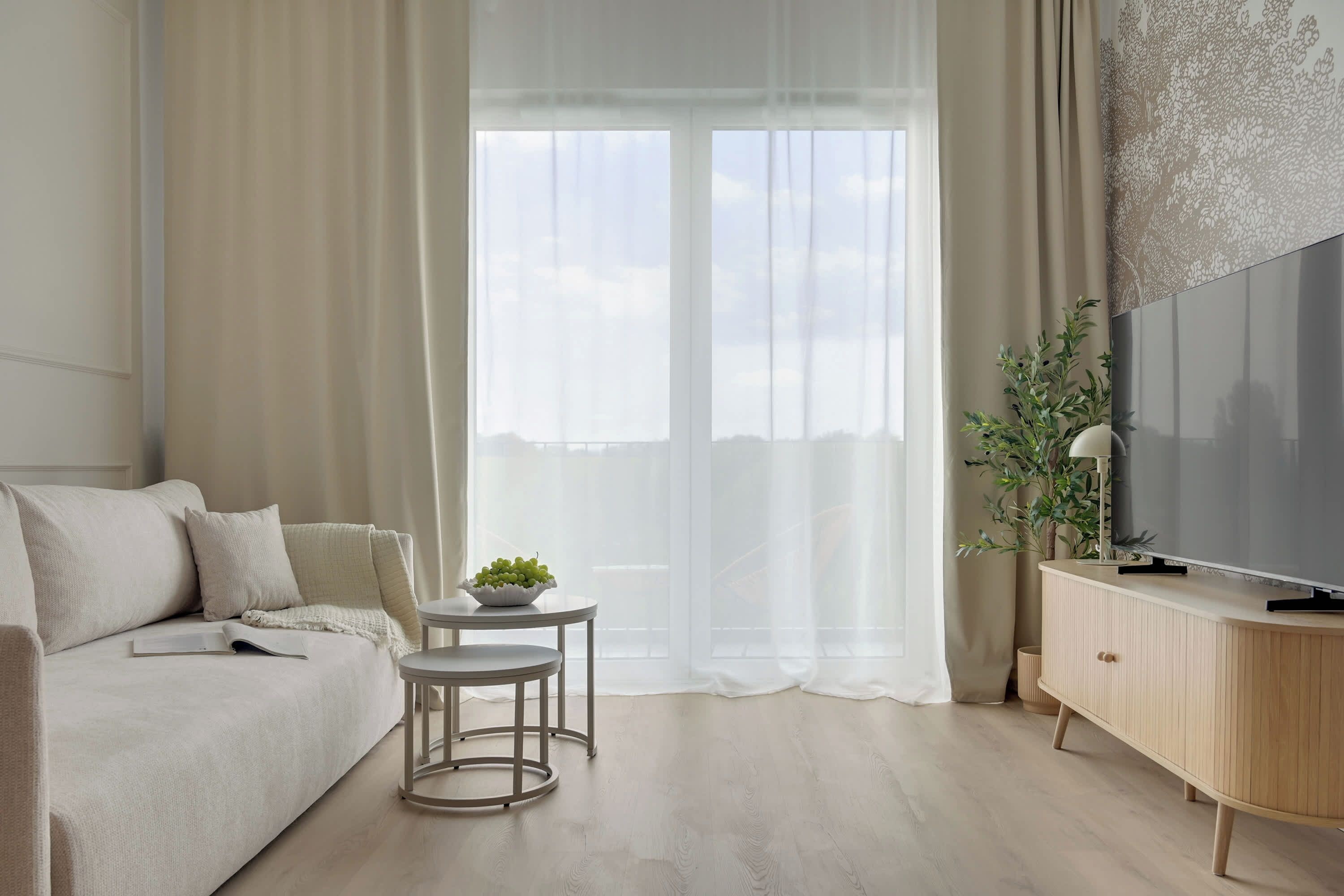 Una sala de estar moderna con un sofá, una mesita redonda con un frutero, un televisor sobre un mueble de madera y cortinas de gasa.