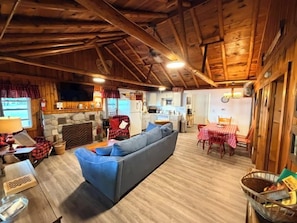 Ein gemütliches Blockhauskaminzimmer mit Kamin, einer Couch, einem Esstisch mit Stühlen und einer Küchenzeile.