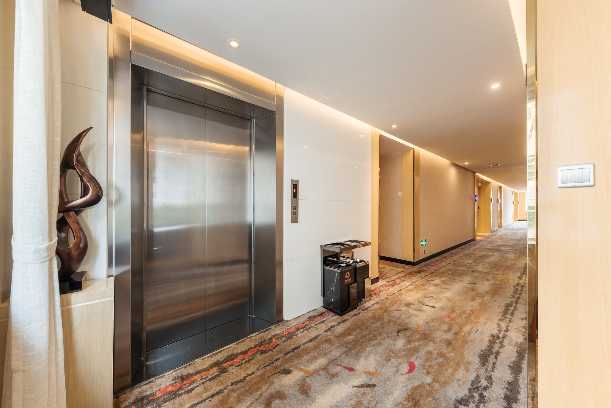 A hotel corridor with a stationary elevator, a trash bin, and a carpeted floor.