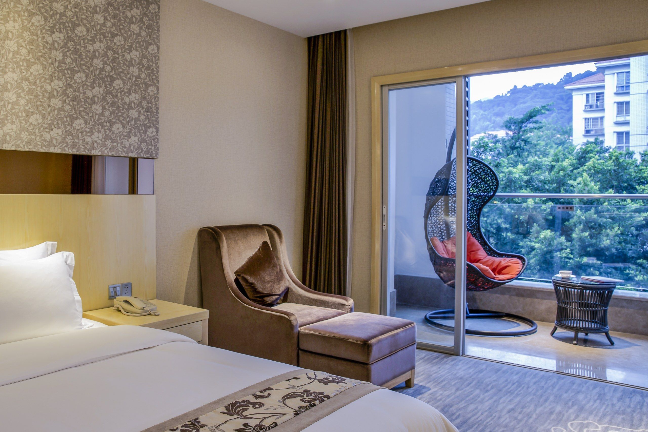 A hotel room with a bed, a chair, a phone, a window with a view of greenery, and a balcony with a chair.