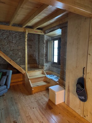 A rustic interior with a wooden staircase leading to a loft area, a stone wall, and a window with a view.