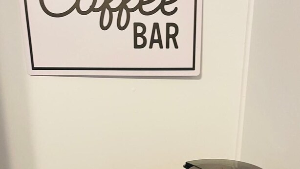 En kaffestation med en kaffebryggare, koppar och en korg placerad på ett träbord framför en vägg med en skylt som visar texten ”Kaffebar”.