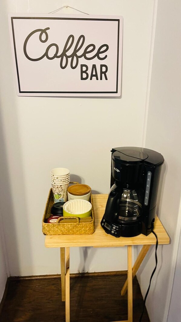 Een koffiebar inrichting met een koffiezetapparaat, kopjes en een mandje op een houten tafel, voor een muur met een 'Coffee Bar' bord.