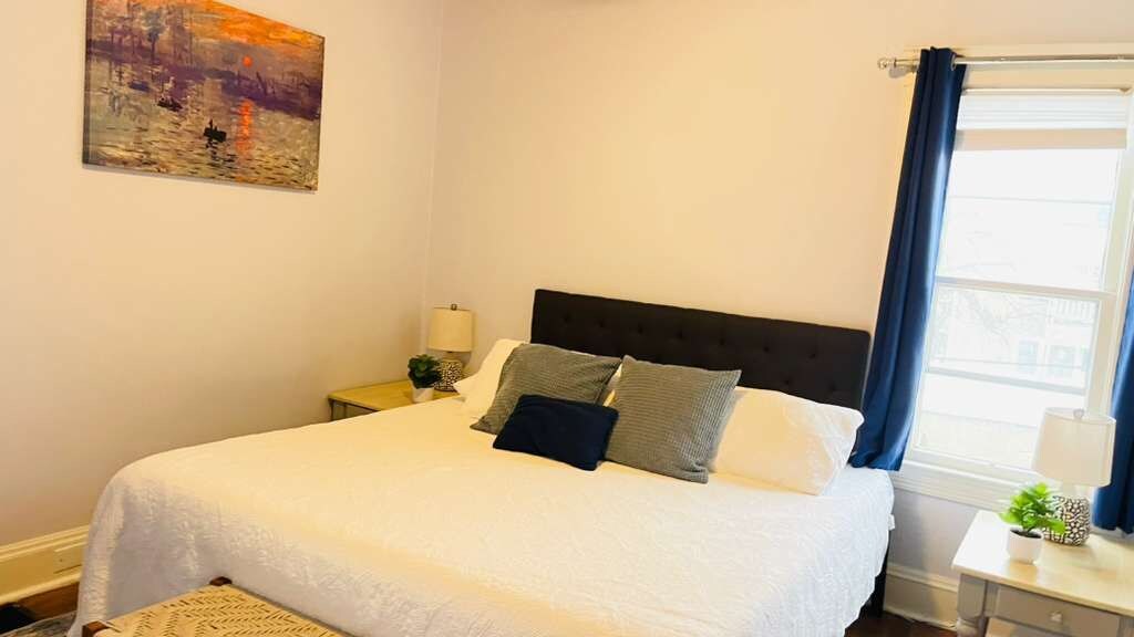 A bedroom with a bed, a nightstand, a lamp, a painting, and a window with curtains.