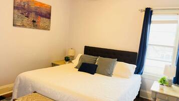A bedroom with a bed, a nightstand, a lamp, a painting, and a window with curtains.