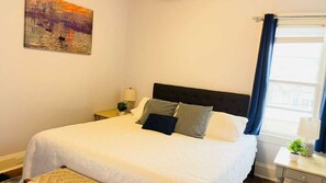 A bedroom with a bed, a nightstand, a lamp, a painting, and a window with curtains.