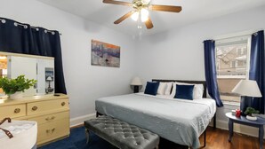Ein Schlafzimmer mit einem Bett, einem Schminktisch, einem Spiegel, einem Deckenventilator, einem Fenster mit Vorhängen und einem Bild an der Wand.