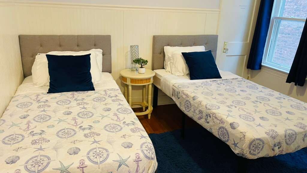A room with two beds, each with a headboard, a small round table with a plant, and a window with curtains.