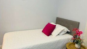 Un lit simple avec une couette blanche, une tête de lit grise et un oreiller rouge. Il y a une petite table de chevet en bois sur laquelle est posé un vase de fleurs.