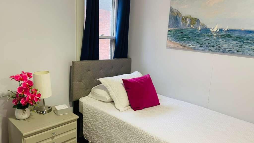 A hotel room with a bed, a nightstand, a lamp, and a painting of a coastal scene.
