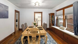 Un comedor con una mesa de madera, sillas, un florero con flores y vista a un edificio de ladrillo a través de una ventana.