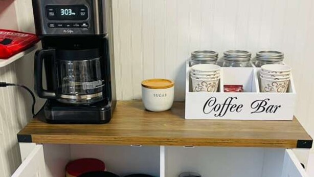 Una barra de café con una cafetera, un recipiente de azúcar y tazas de café sobre una estantería, y contenedores de almacenamiento en la repisa inferior.