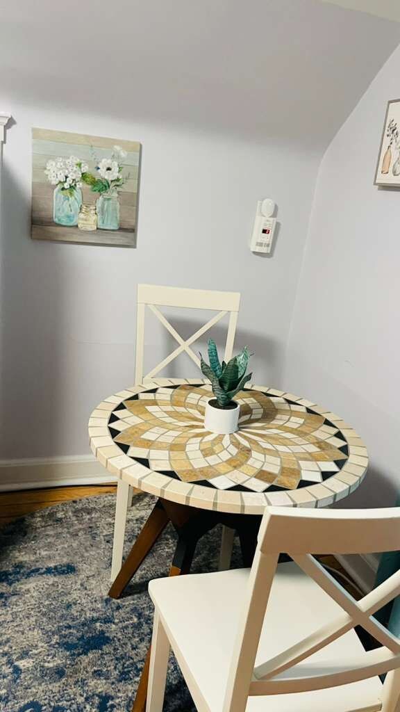 A round table with a mosaic top, two white chairs, a potted plant, and a painting on the wall.