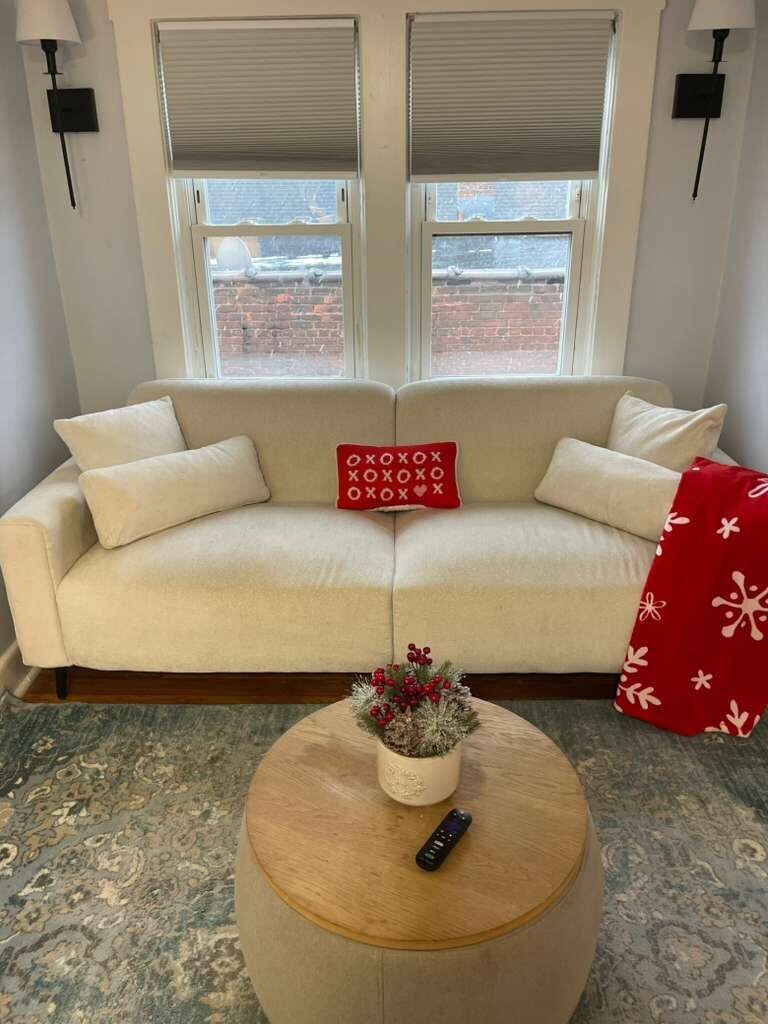 A beige sofa with a red cushion, a small round wooden table with a remote control, and a window with blinds.