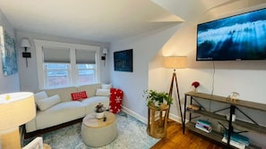 Een woonkamer met een bank, een salontafel, een aan de muur bevestigde televisie en een raam met zonwering.