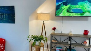 Een kamer met een aan de muur bevestigde televisie, een lamp, een plant en een plank met boeken en een rode mok.