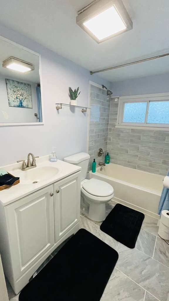 Ein Badezimmer mit einem weißen Waschbecken, einer Toilette und einer Badewanne. Über der Badewanne befindet sich ein Fenster, über dem Waschbecken ein Spiegel.