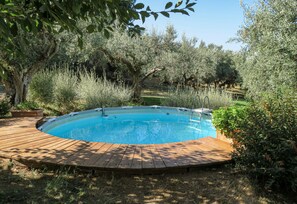 Una piscina circular con una terraza de madera alrededor, ubicada en un jardín con diversos árboles y arbustos.