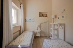 A bedroom with two single beds, a bedside table with a lamp, a radiator, a wardrobe, and a small shelf with books and pictures.