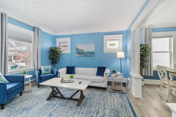 Een woonkamer met een witte bank, blauwe fauteuils, een salontafel en een kamerkleed met een patroon.