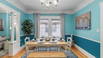 Uma sala de jantar com mesa de madeira posta para quatro, cadeiras brancas, um lustre e uma janela grande com cortinas.