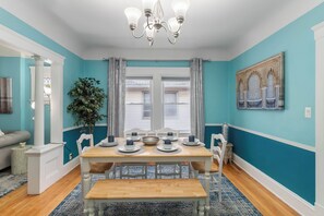 A dining room with a wooden table set for four, white chairs, a chandelier, and a large window with curtains. - ✨ Family-Friendly Charm in the Heart of Lakewood 🌿<br>Steps from Parks, Trails & Lake Views<br>Welcome to your serene escape in Lakewood—where convenience meets charm in one of Cleveland’s most walkable neighborhoods. (Lakewood)
