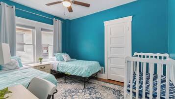 A bedroom with two beds, a crib, a ceiling fan, and a window with curtains.