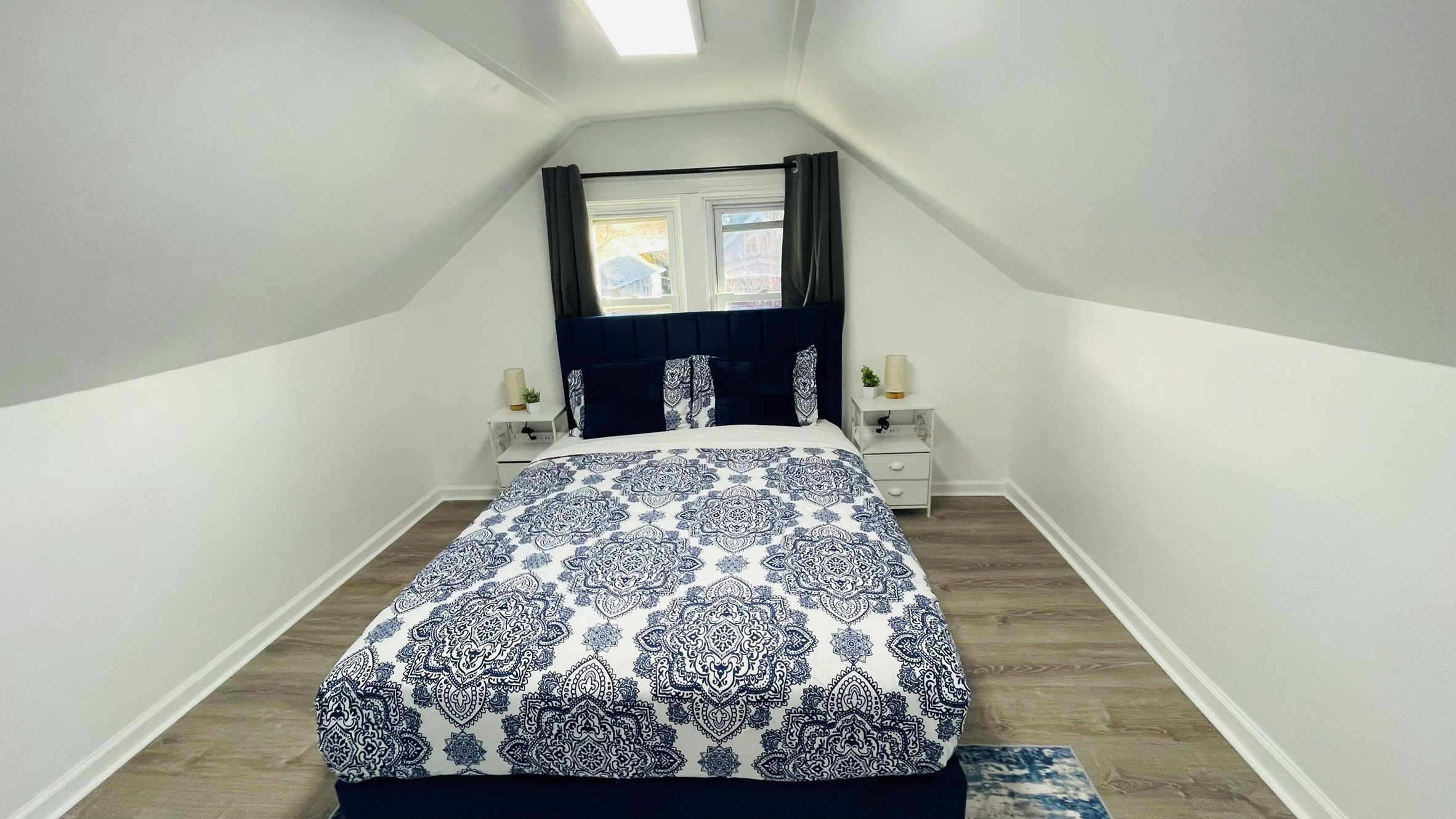 A narrow, sloped room with a bed centered, flanked by two bedside tables with lamps, a window with curtains, and a skylight.