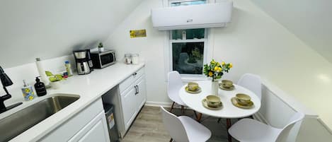 A small kitchenette with a microwave, coffee maker, and sink. There is a dining area with a table set for two and a vase with flowers.