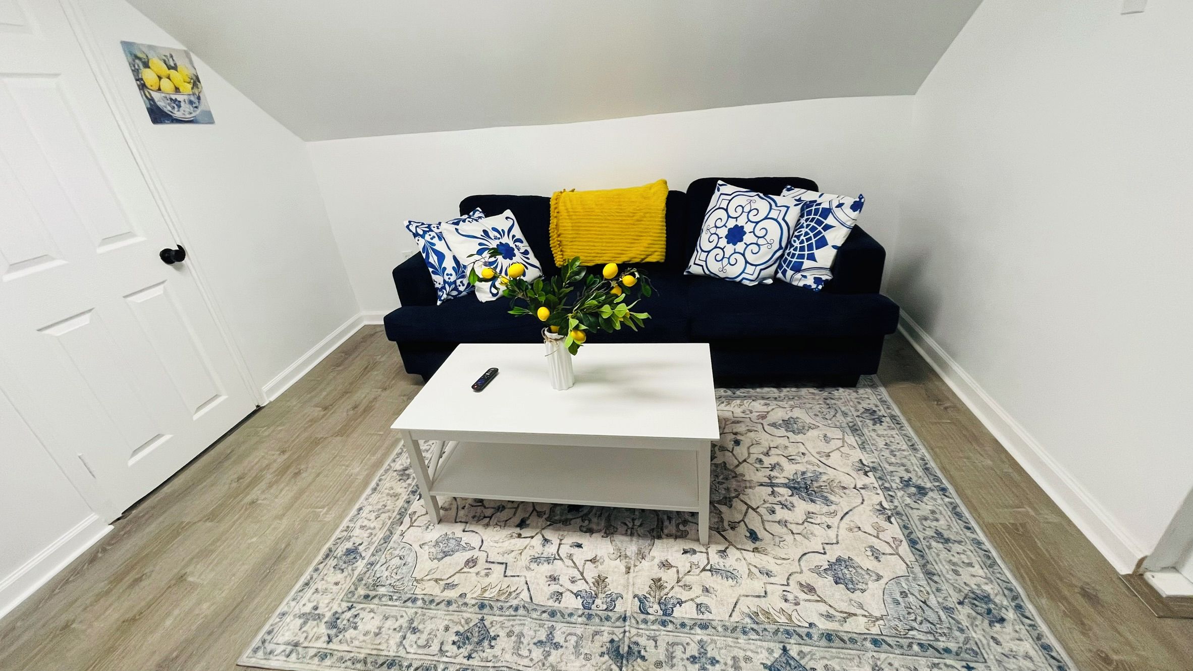 Un salon avec une table basse blanche, un tapis à motifs bleus et blancs, un canapé bleu foncé agrémenté de coussins jaunes et à motifs, et un vase rempli de fleurs jaunes.