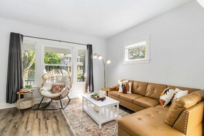 Un salon avec un canapé en cuir marron, un fauteuil en osier, une table basse blanche et une grande fenêtre avec des rideaux.