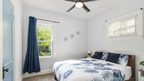 Ein Schlafzimmer mit einem Bett, einem Deckenventilator, einem Fenster mit Vorhängen und einem kleinen Nachttisch mit Lampe.