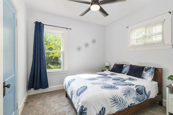 Ein Schlafzimmer mit einem Bett, einem Deckenventilator, einem Fenster mit Vorhängen und einem kleinen Nachttisch mit Lampe.