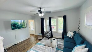A living room with a flat-screen TV, a ceiling fan, a sofa, a coffee table, and a view of the outdoors through large windows. - Your Coastal Getaway in West Cleveland! (Cleveland)