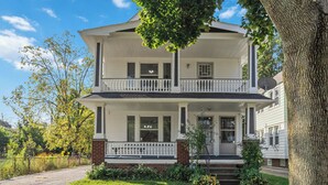 Ein zweistöckiges Haus mit einer Veranda, einem Ziegelkeller und einem großen Baum im Vordergrund.