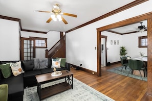Ein geräumiges Wohnzimmer mit Holzboden, einer schwarzen Couch mit grünen Kissen, einem Couchtisch und einem Deckenventilator.