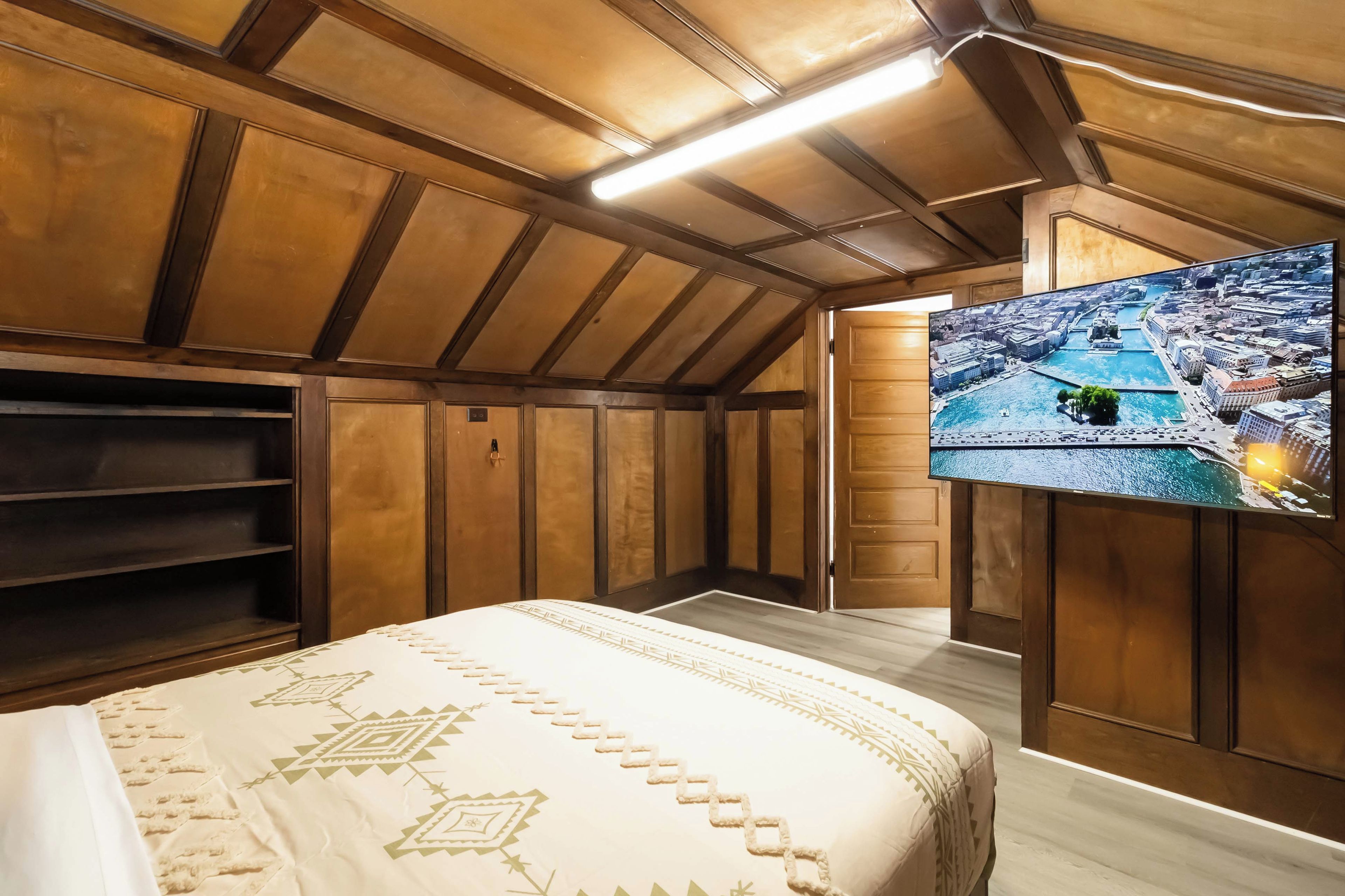 Una habitación con cama, paneles de madera, televisor mostrando un paisaje urbano y techo inclinado.
